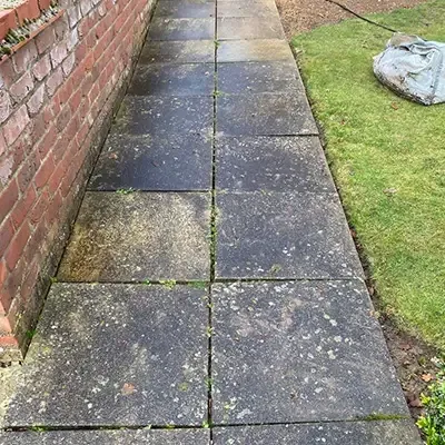 Patio slabs beside a brick wall, dark with algae before cleaning